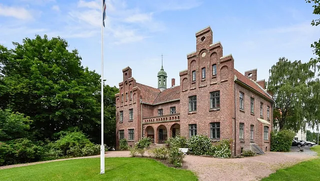 Mursteinsbygning med trappegavler, buet inngangsparti og flaggstang foran på en grønn plen.
