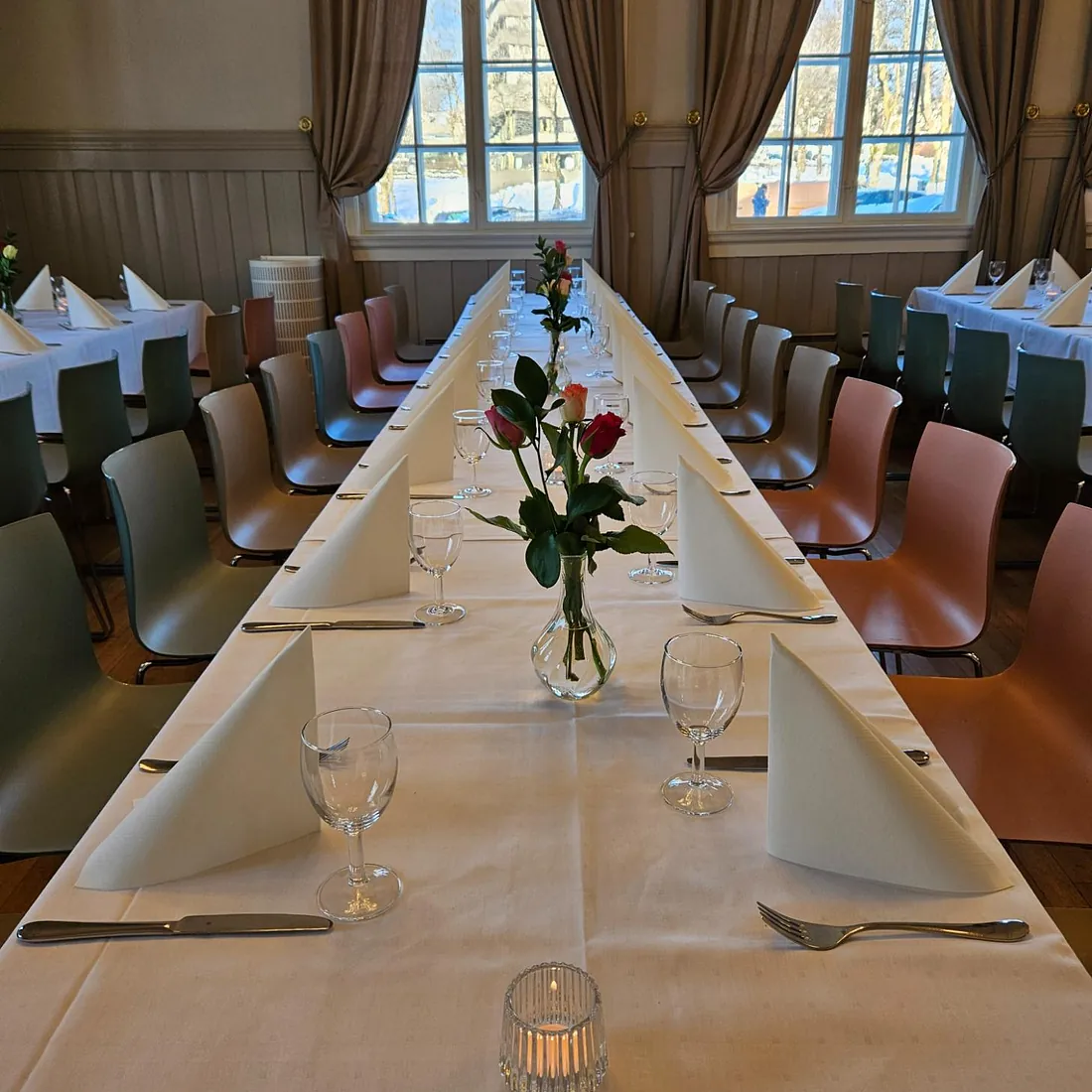 Langbord med hvit duk og roser i vase, foran høye vinduer med gardiner og vinterutsikt.