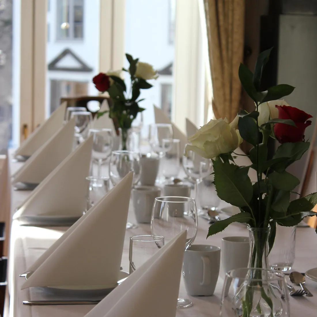 Hvitdekket bord med pyntede glass og roser i vase, foran lyse vinduer med gardiner.