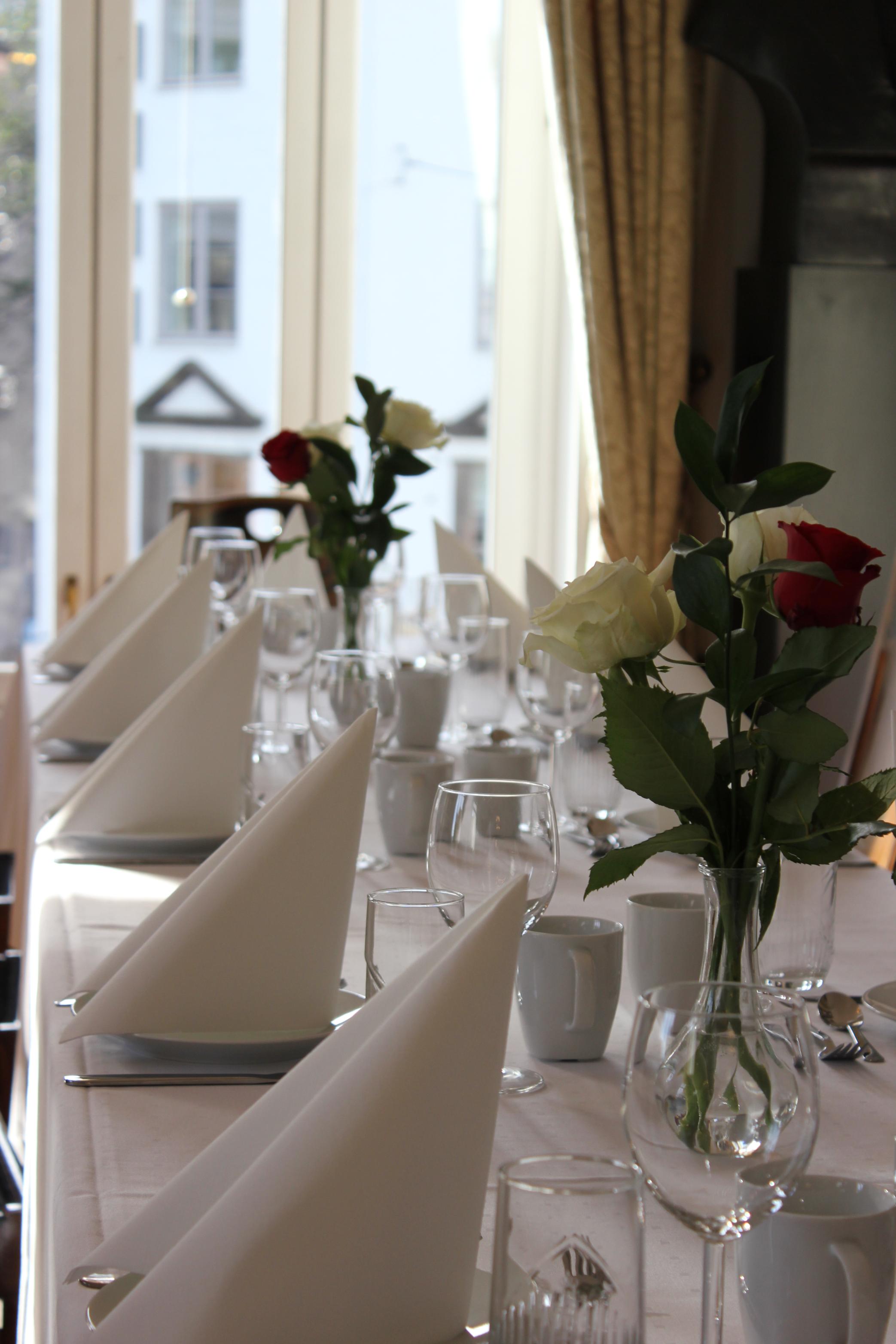 Hvitdekket bord med pyntede glass og roser i vase, foran lyse vinduer med gardiner.
